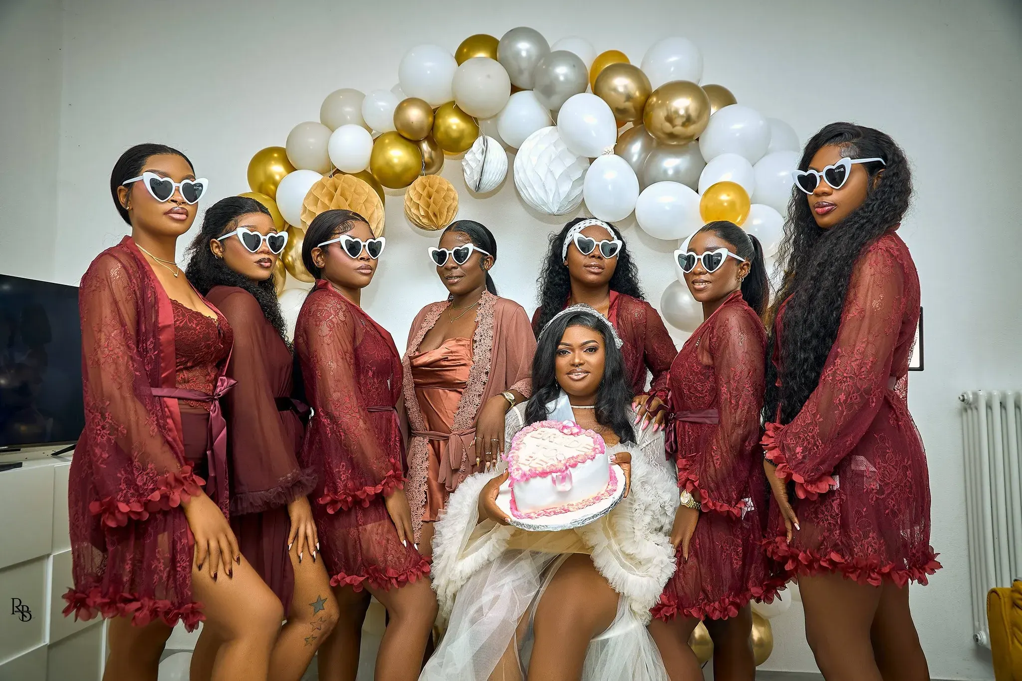 Bride holds a cake while bridesmaids in burgundy robes stand around her.