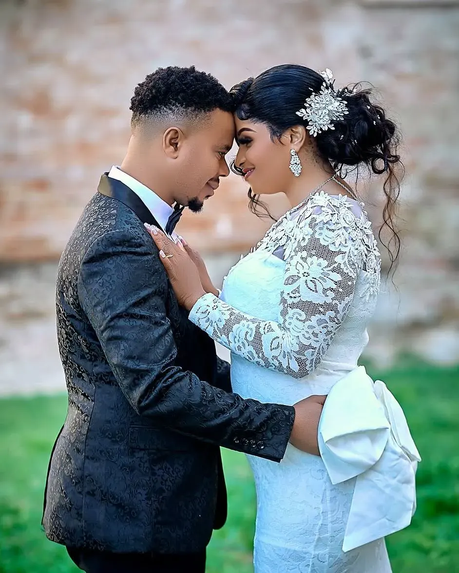 Couple touching foreheads in front of a brick wall, the bride in a lace gown and the groom in a black jacquard suit.